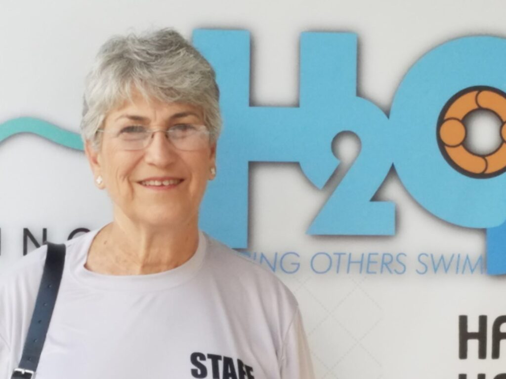 A woman with short grey hair and glasses is smiling, standing in front of a blue and orange logo that reads "H2O" and "ING OTHERS SWIM."