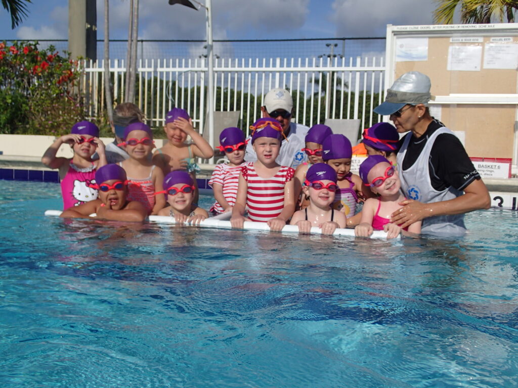 A group of children in swim caps and goggles standing in a pool with an adult instructor.