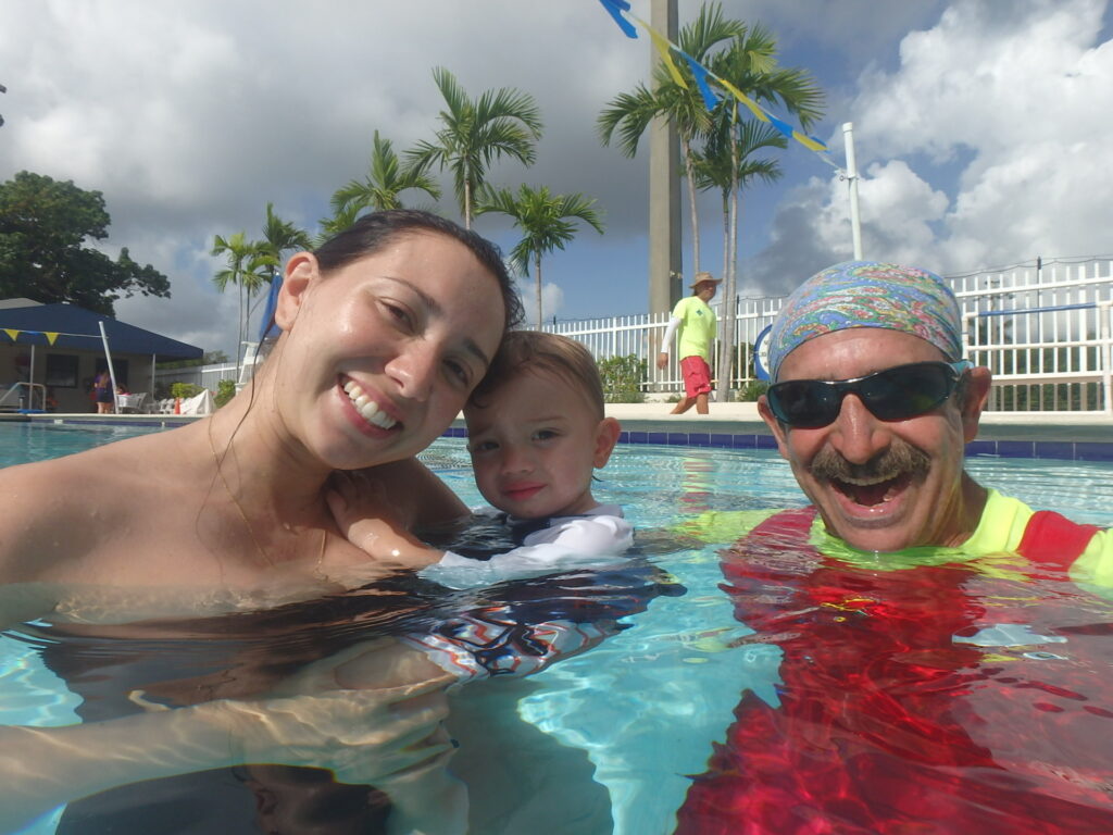 A woman and a man are in a swimming pool with a young child, all smiling at the camera.