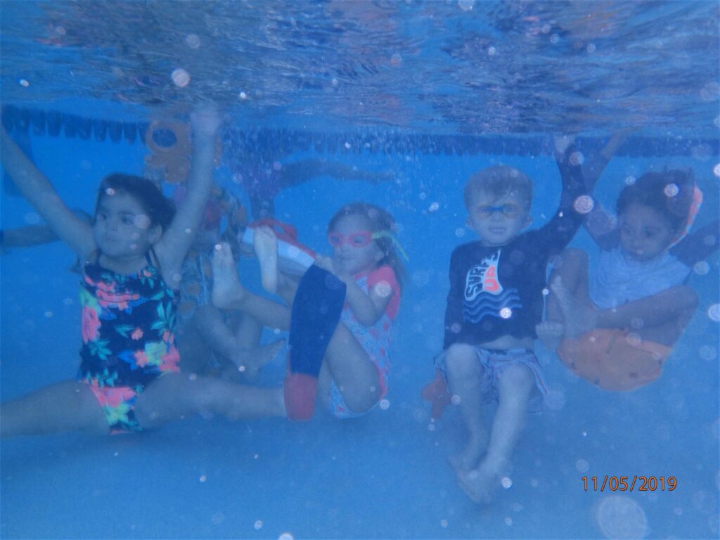 Several children are seen underwater in a swimming pool, some with goggles, smiling.