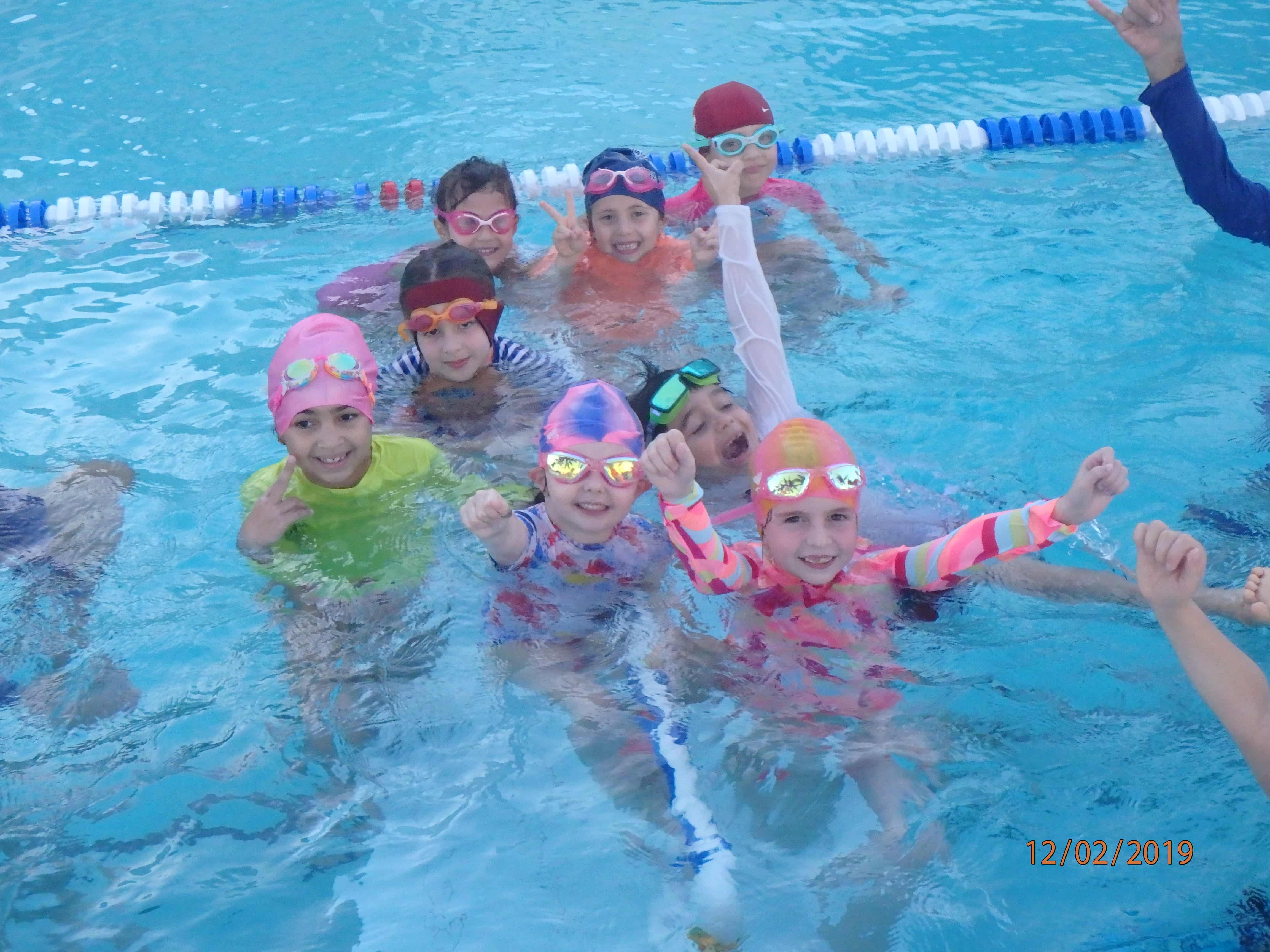 A group of children in swim caps and goggles smiling and posing in a swimming pool.